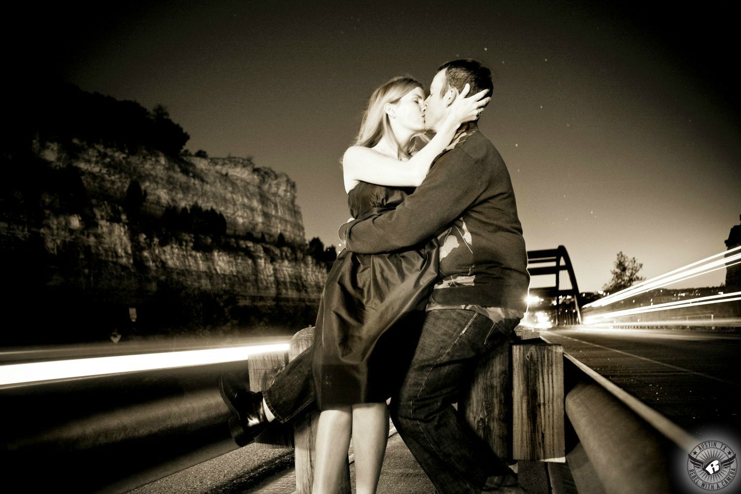 Passionate blond wearing a dark knee length dress kisses a dark haired guy with a long sleeve dark sweater and blue jeans between the guard rails in the median on 360 Highway in front of the Pennybacker Bridge  at night with car headlights and taillights streaming by at night in this long exposure engagement portrait in Austin, Texas.  